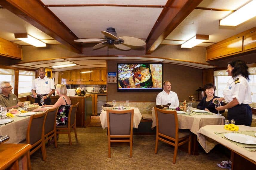 Dining room of Bahamas Aggressor, guests eating, a fan on the ceiling and chairs in view. Has a brown background from decor.